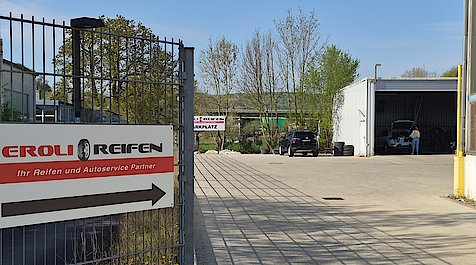 Das Bild zeigt die Einfahrt zu „EROLI REIFEN“ mit Hinweisschild, Hof und geöffneter Werkstatthalle mit Autos.