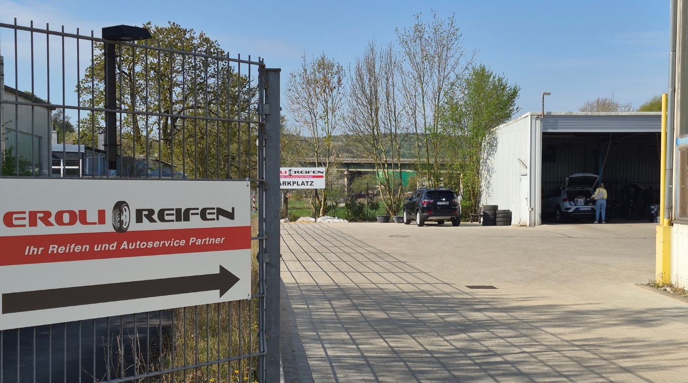 Das Bild zeigt die Einfahrt zu „EROLI REIFEN“ mit Hinweisschild, Hof und geöffneter Werkstatthalle mit Autos.