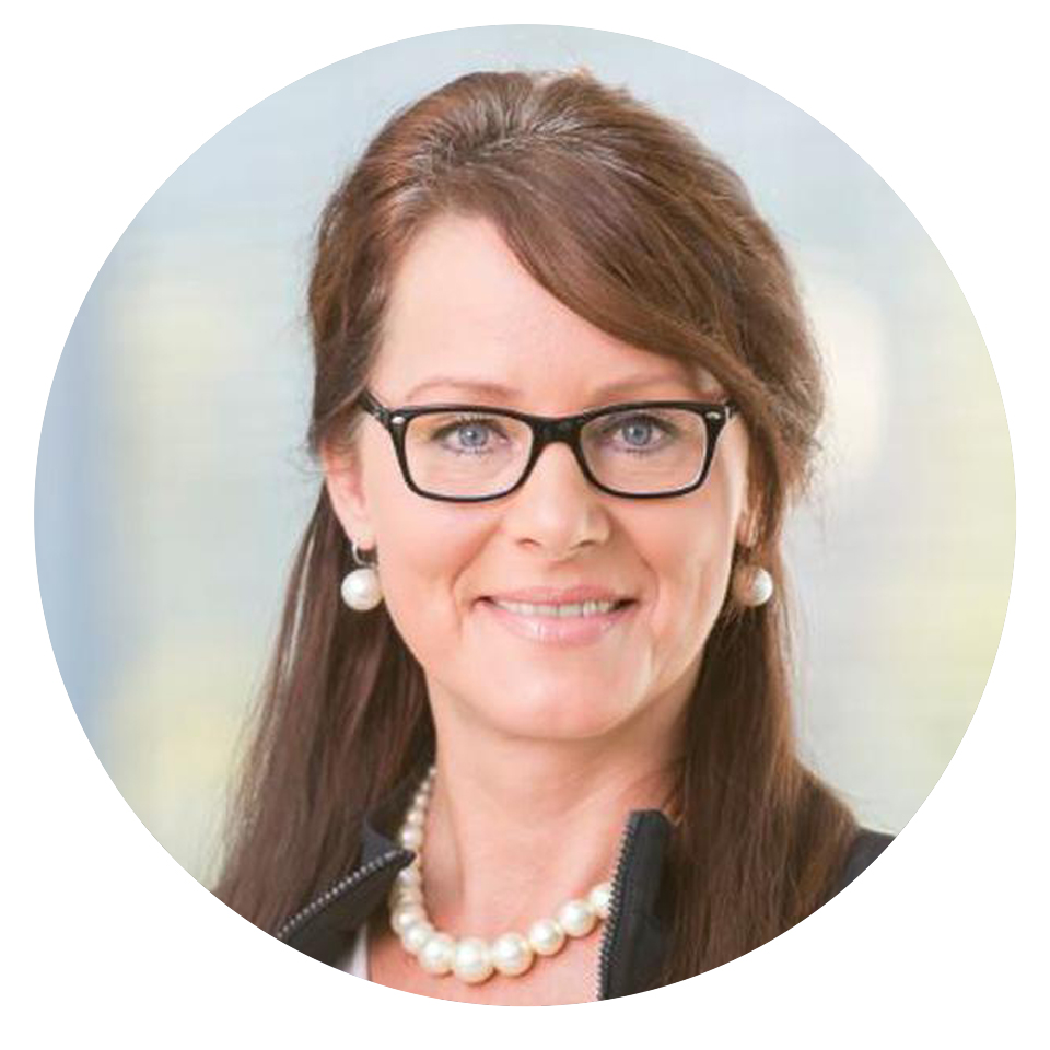 Frau mittleren Alters mit Brille und Perlenkette lächelt freundlich in einem professionellen Porträt.