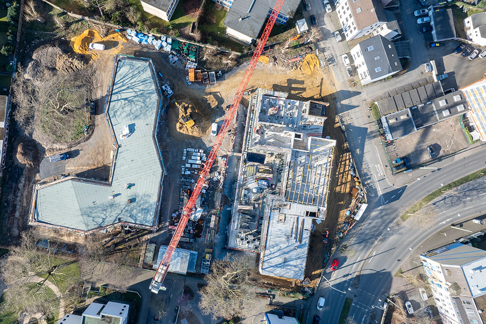 Blick aus der Vogelperspektive auf eine Baustelle: Angrenzend an eine Straße entsteht ein neues Gebäude, man sieht Baustellenfahrzeuge und einen Kran.