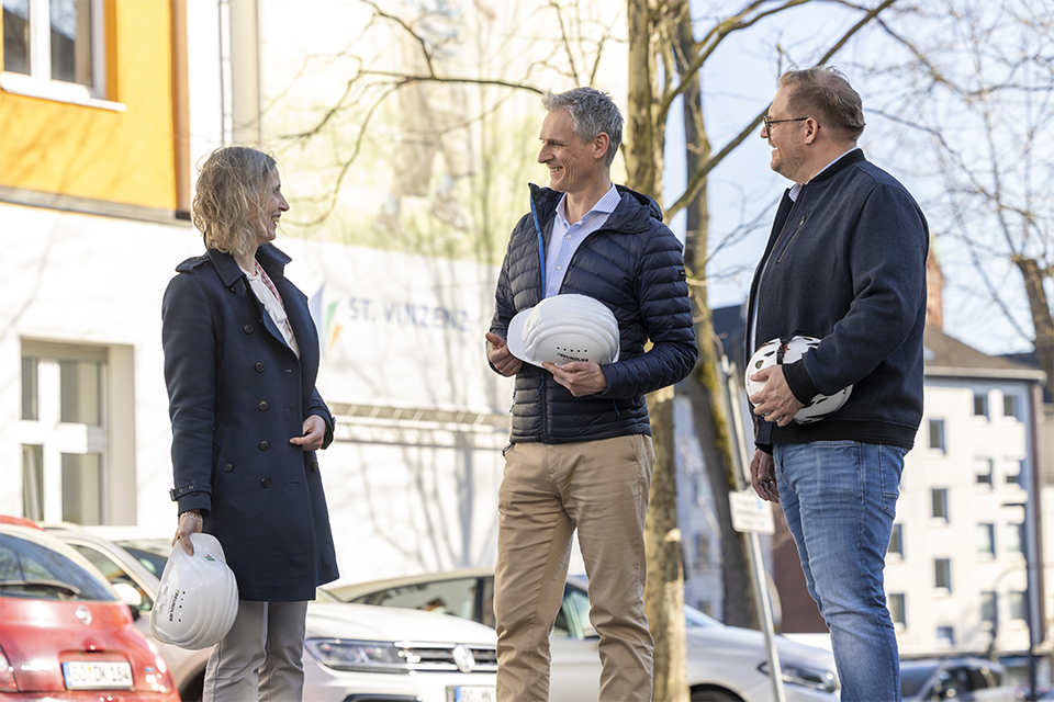 Drei Personen stehen vor einem Gebäude und unterhalten sich. Sie haben jeder einen Bauhelm in der Hand. An dem Gebäude im Hintergrund hängt ein Schild mit der Aufschrift "ST. VINZENZ".