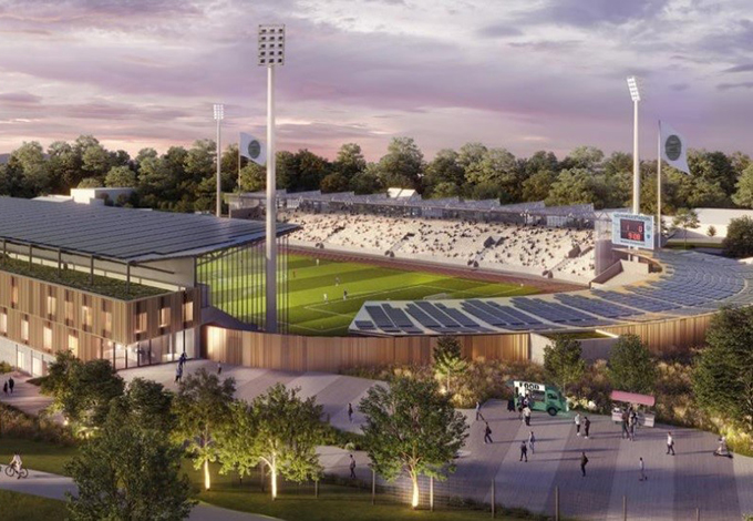 Modernes Fußballstadion mit Spielfeld, leeren Rängen, Flutlichtmasten und umgeben von Bäumen.
