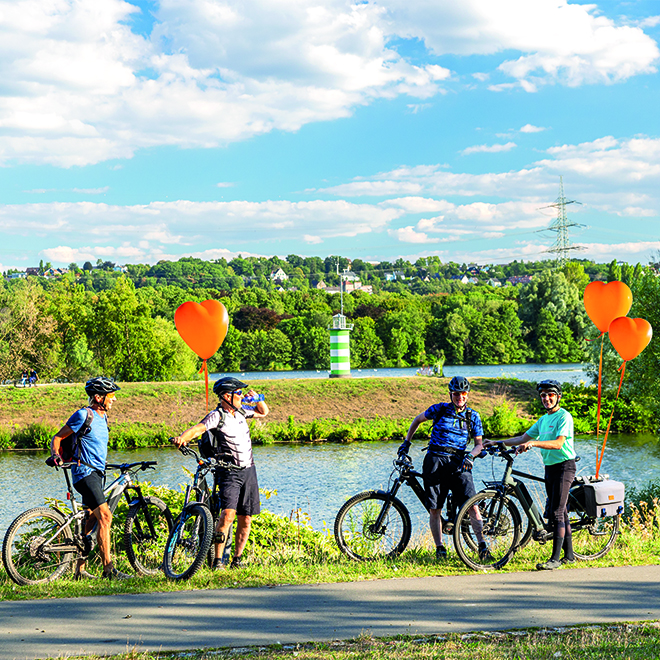 Vier Radfahrer mit Helmen pausieren an einem Fluss, während sie leuchtend orangefarbene Ballons halten.
