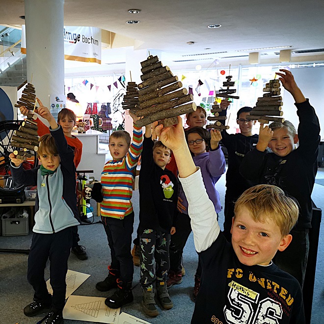 Kinder halten selbstgebastelte Mini-Weihnachtsbäume aus Holzästen stolz in die Höhe, drinnen in Gruppen.