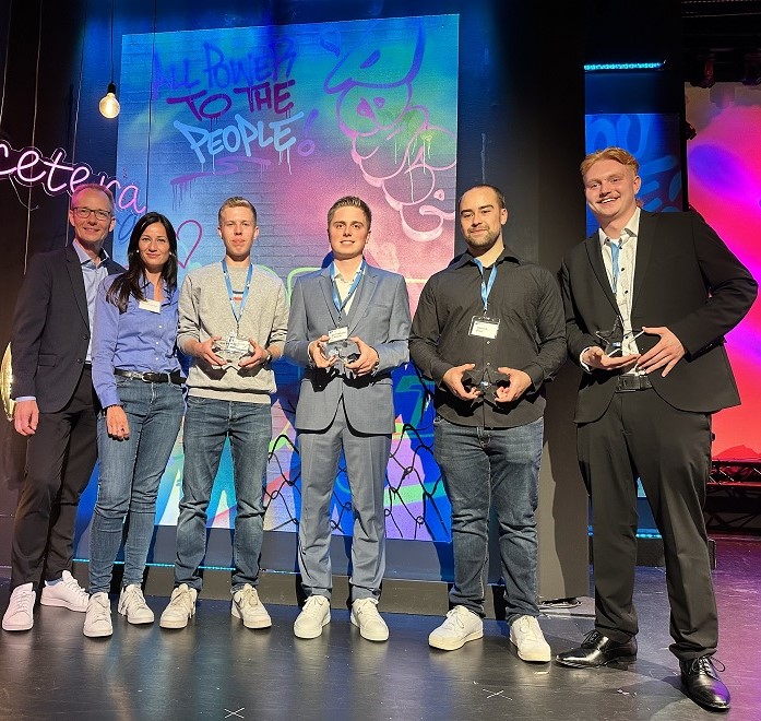 Sechs Personen auf einer Bühne, darunter vier Preisträger mit Trophäen in den Händen, und zwei Moderatoren.