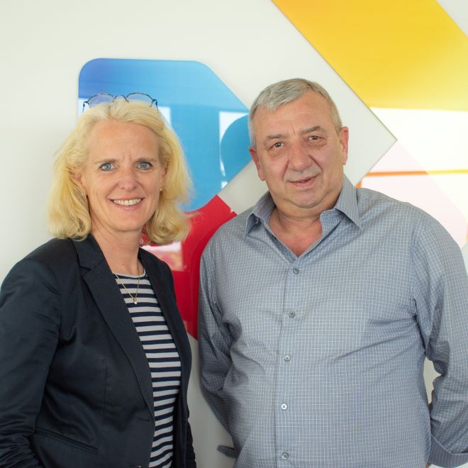 Zwei Geschäftsleute stehen vor einer Wand mit geometrischen Mustern und lächeln in die Kamera.