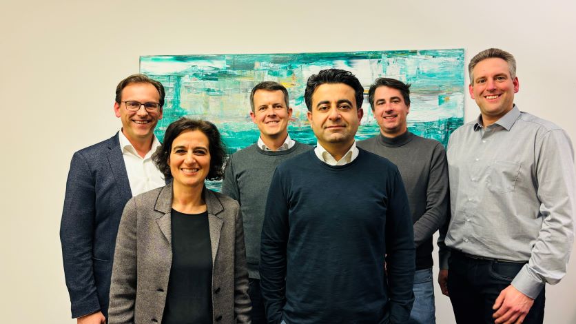 Gruppe von sechs lächelnden Personen, drei Frauen, drei Männer, die in einem Büro vor einem Gemälde stehen.