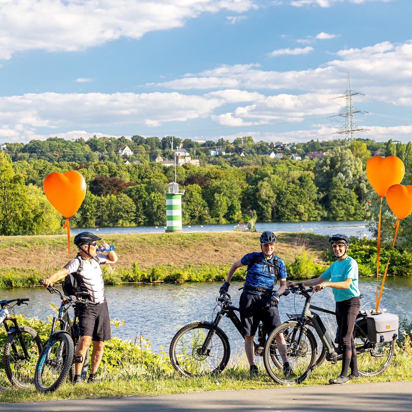 Gruppe von drei Radfahrern mit Luftballons posiert lächelnd an einem Flussufer bei sonnigem Wetter.