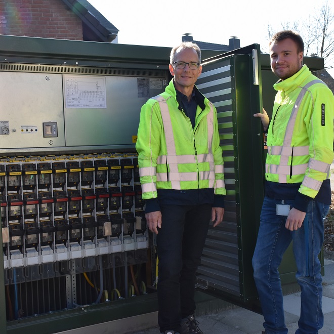 Zwei Männer in Schutzjacken stehen vor einem geöffneten Schaltschrank und schauen in die Kamera.