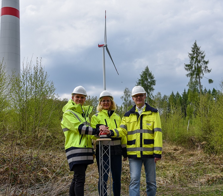 Drei Personen in Schutzkleidung stehen vor einem Windrad und halten gemeinsam feierlich einen Knopf.