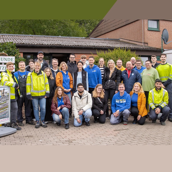 Gruppenfoto von 28 fröhlich lächelnden Menschen in Arbeitskleidung vor einem Gemeindegebäude.