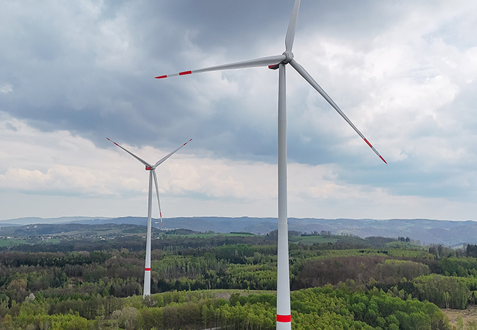 Zwei Windräder vor bewölktem Himmel und Wald