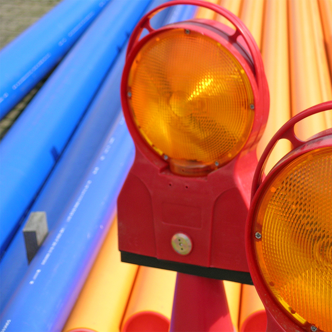 Straßenbaulampen stehen vor gestapelten Kunststoffrohren auf einer Baustelle in vermutlich urbanem Umfeld.