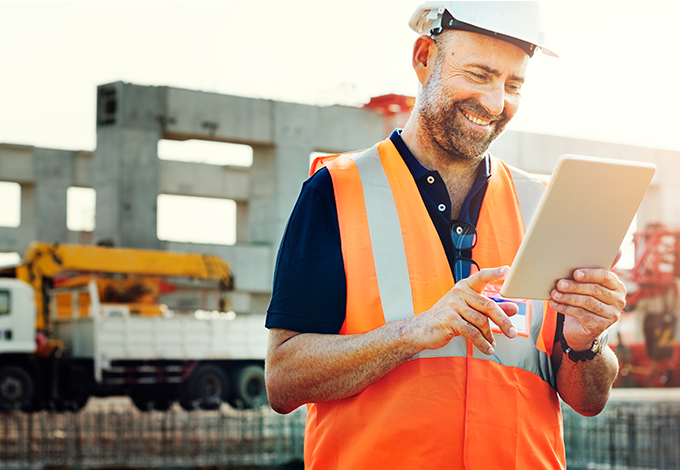 Bauarbeiter mit Schutzhelm und Warnweste nutzt ein Tablet auf einer Baustelle mit Kran im Hintergrund.