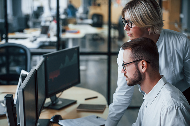 Zwei Businesspersonen im Büro vor mehreren Computermonitoren.