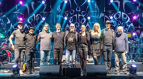 Gruppe von Musikern auf einer Bühne mit Lichteffekten und Instrumenten im Hintergrund.