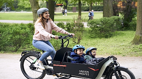 Lächelnde Frau mit Helm mit Lastenrad und 2 Kindern mit Helm