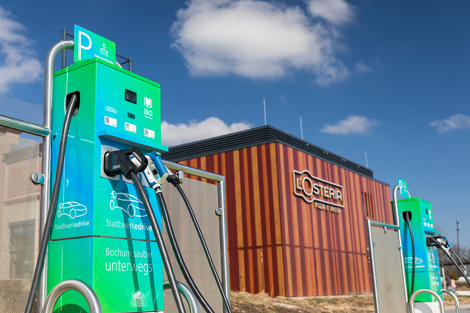 Ladestation mit mehreren Ladepunkten vor einem Gebäude mit "L'Osteria"-Logo im Hintergrund.