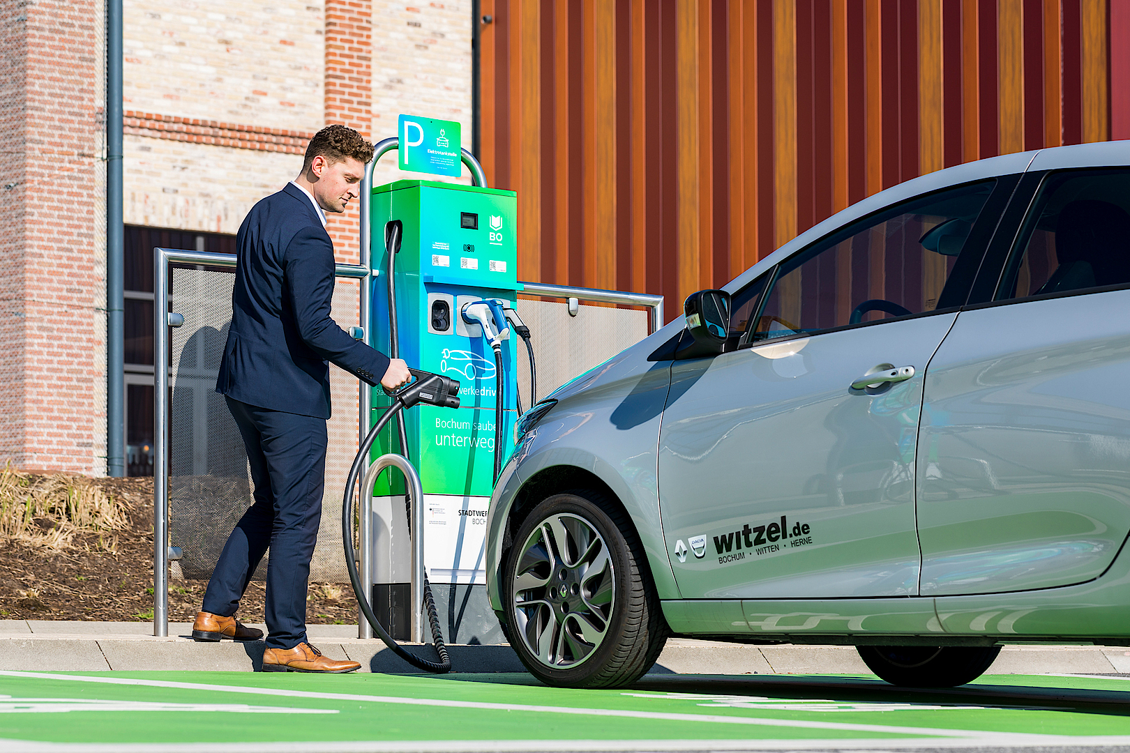 Mann lädt Elektroauto an Ladestation auf, modernem Gebäude im Hintergrund.