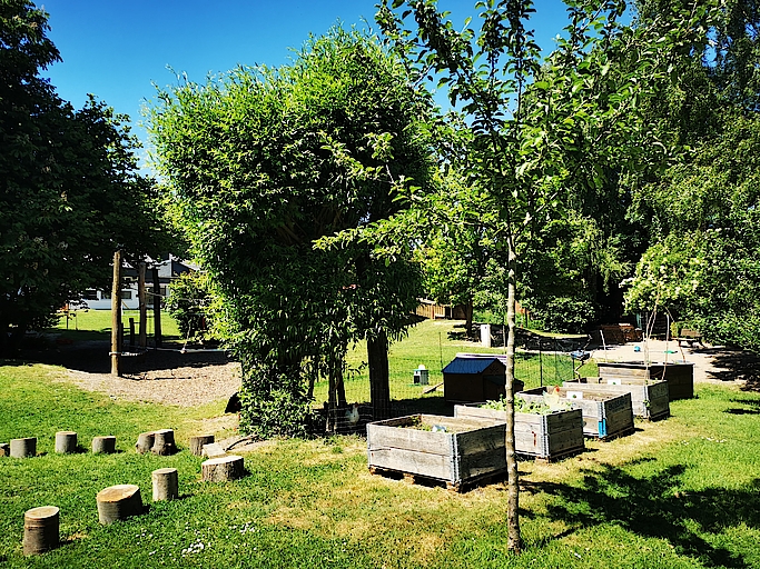 Grüner Spielplatz mit Hochbeeten und Bäumen im Sonnenlicht