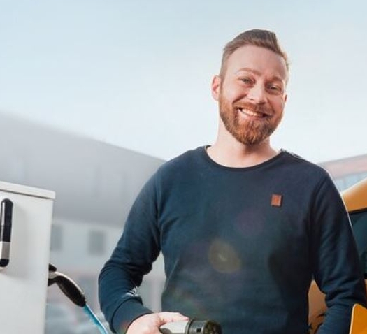 Lächelnder Mann lädt sein Elektroauto an einer Ladestation auf, im Hintergrund ein modernes Gebäude.