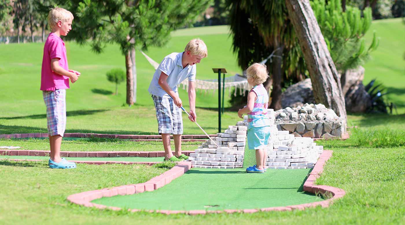 Kinder spielen Minigolf auf einer grünen Anlage mit Bäumen im Hintergrund.