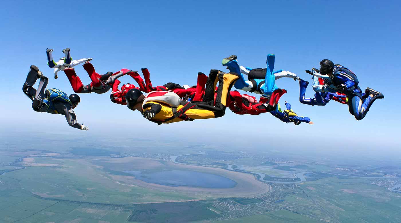 Gruppe von Fallschirmspringern in Formation am Himmel über einer Landschaft.