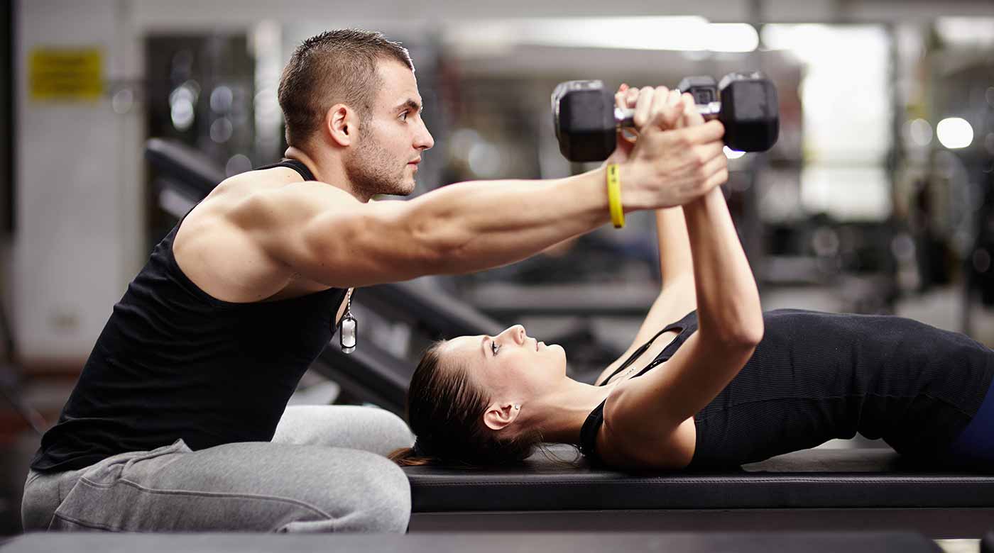 Trainer unterstützt Frau beim Hanteltraining auf einer Bank im Fitnessstudio.