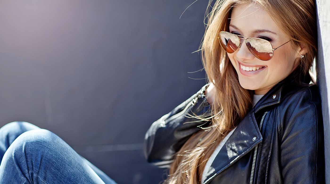 Frau trägt stylische Sonnenbrille, Lederjacke und Jeans, lehnt entspannt an einer Wand.