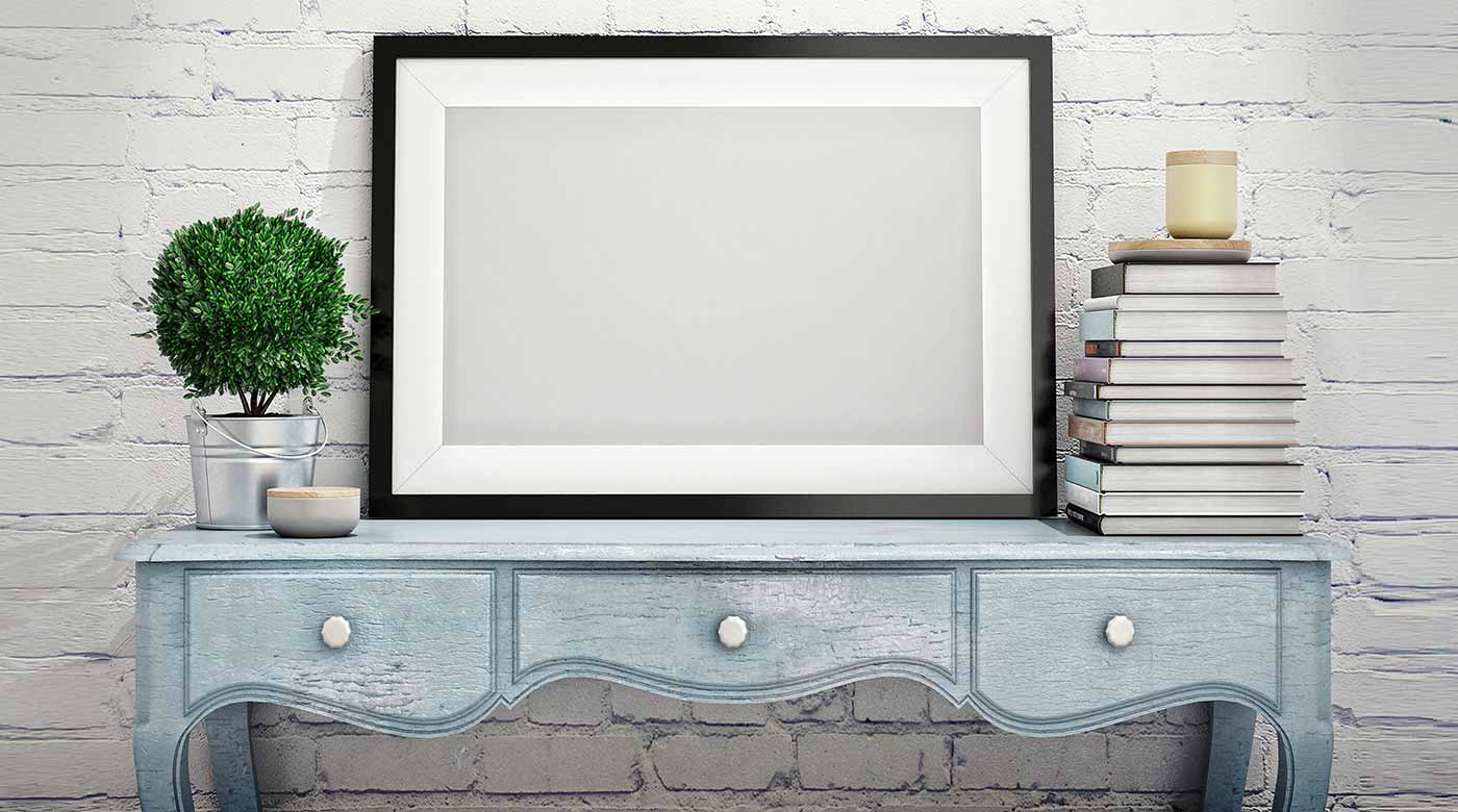 Leerer Bilderrahmen auf blauem Tisch mit Pflanze, Büchern und Kerze vor Ziegelwand.