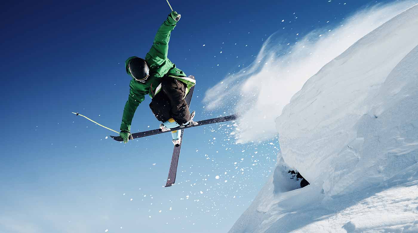 Skifahrer in grüner Jacke springt dynamisch über eine Schneekante vor blauem Himmel.