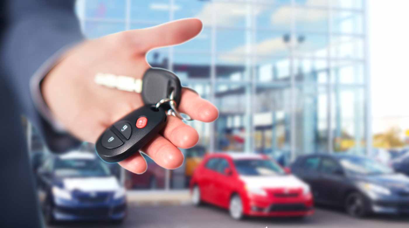 Hand hält Autoschlüssel vor einem Autohaus mit geparkten Fahrzeugen im Hintergrund.