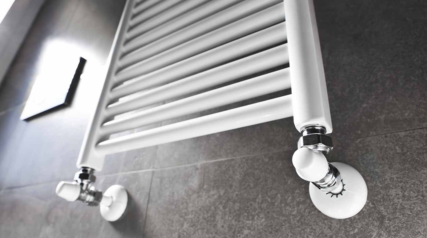 Weißer Handtuchheizkörper mit modernen Anschlüssen vor einer grauen Fliesenwand.
