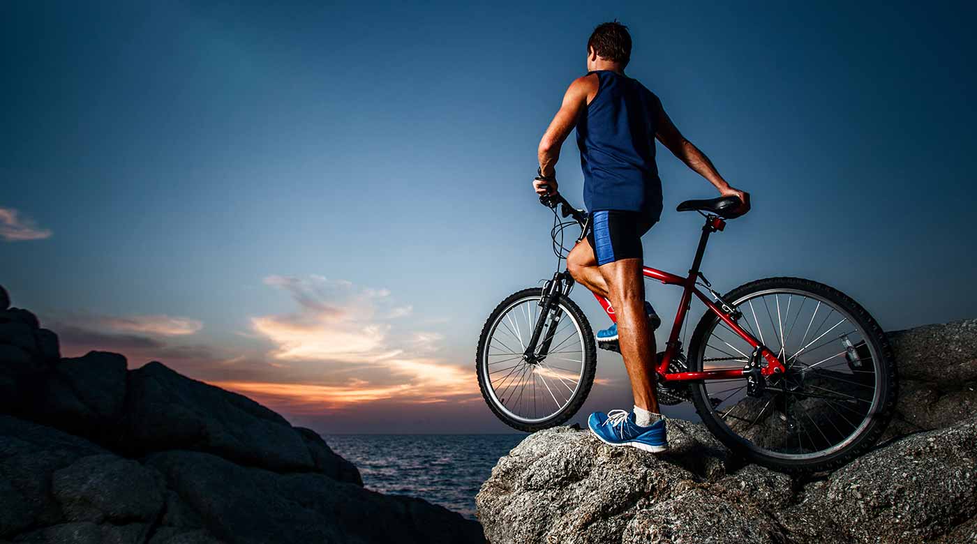 Radfahrer mit rotem Fahrrad steht auf Felsen und blickt auf das Meer bei Sonnenuntergang.