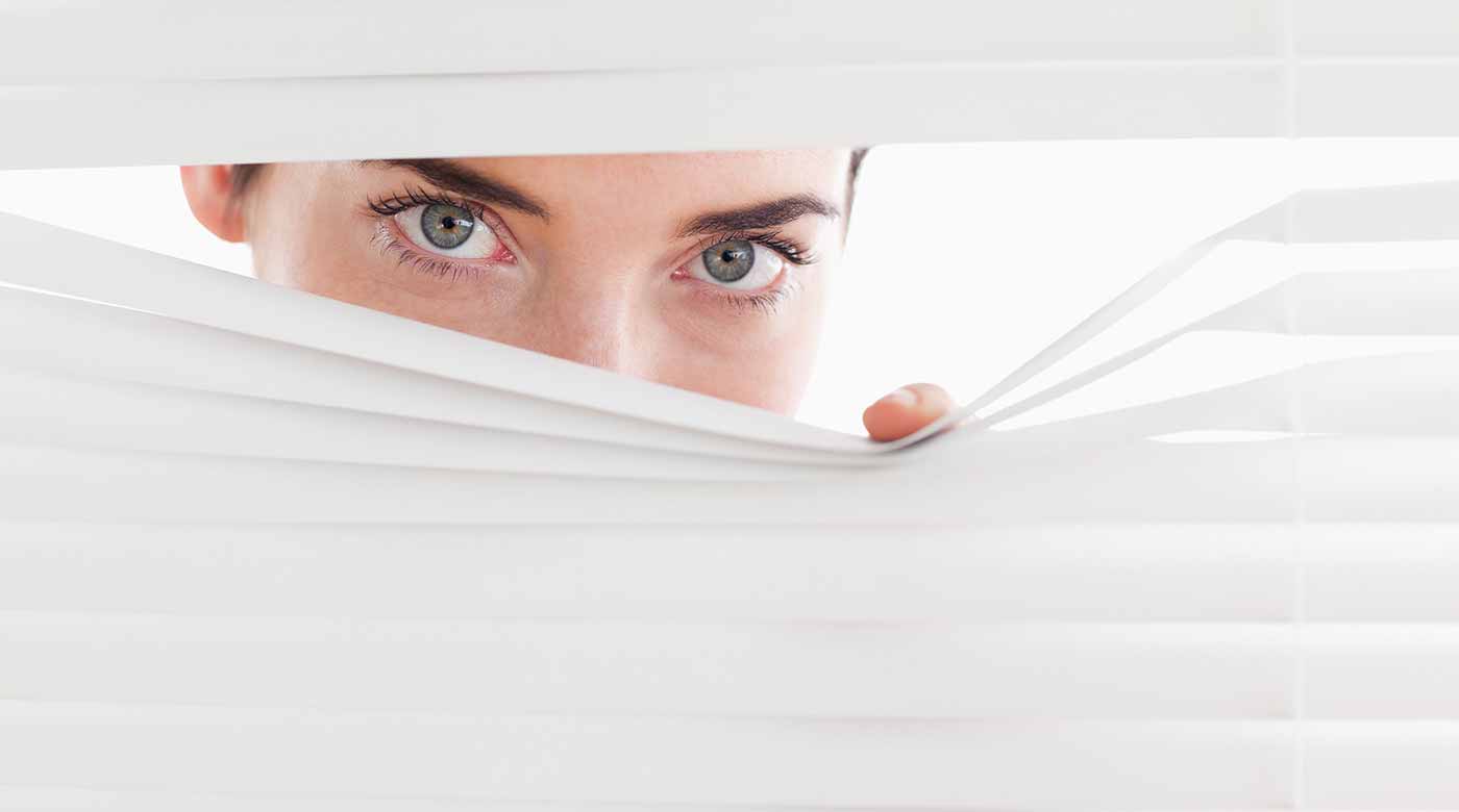 Hand öffnet weiße horizontale Jalousien, um durch die Lamellen ins helle Tageslicht zu sehen.