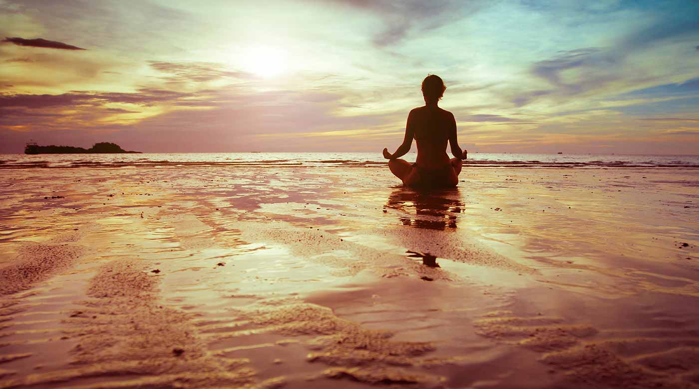 Person meditiert im Lotussitz am Strand bei Sonnenuntergang, ruhige und friedliche Szene.
