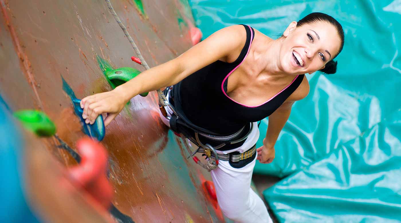 Frau klettert an einer bunten Kletterwand mit Sicherheitsgurt, Matte im Hintergrund.