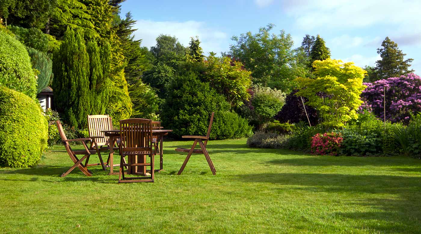 Grüner Garten mit Holz-Gartenmöbeln, bunten Blumen und dichtem Baumbestand.