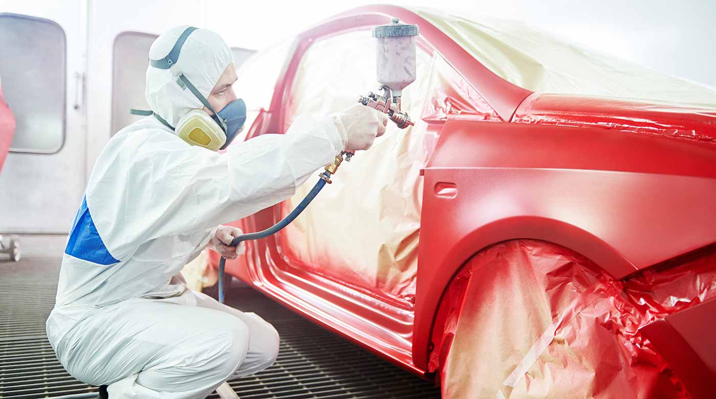 Lackierer in Schutzanzug sprüht rote Farbe auf ein abgeklemmtes Auto in einer Werkstatt.