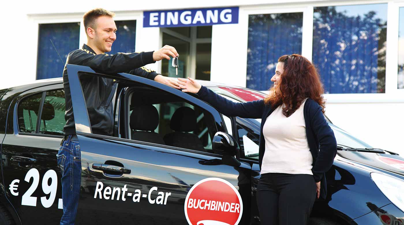 Mitarbeiter übergibt Autoschlüssel an Kundin vor einem Buchbinder-Mietwagen.