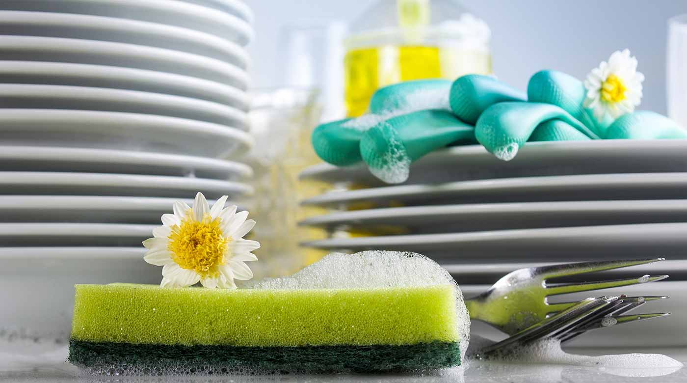 Gestapelte Teller, Schwamm mit Blume, Gabeln, Handschuhe und Spülmittel im Hintergrund.