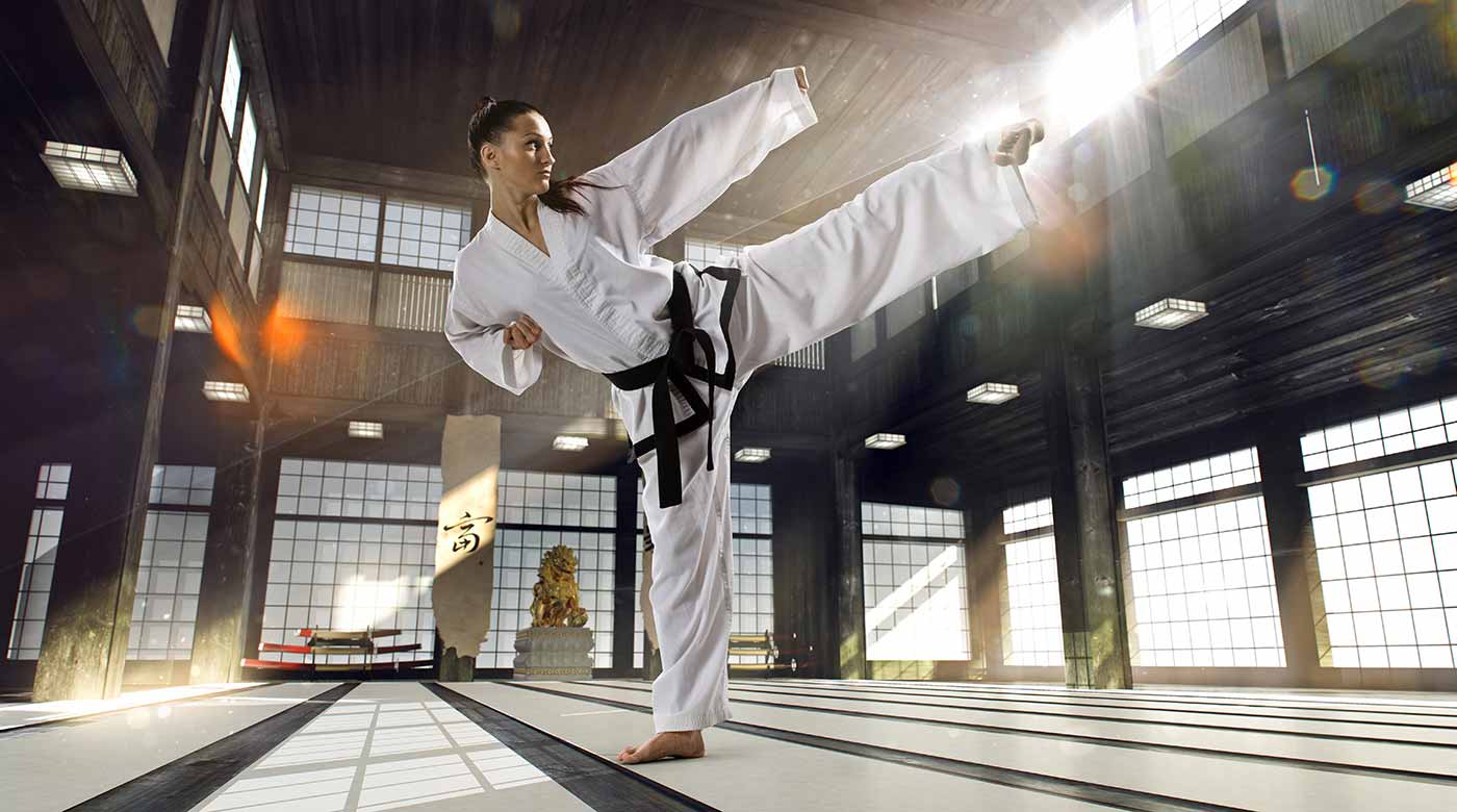 Kampfsportlerin in weißem Anzug führt hohen Kick in traditioneller Halle aus.