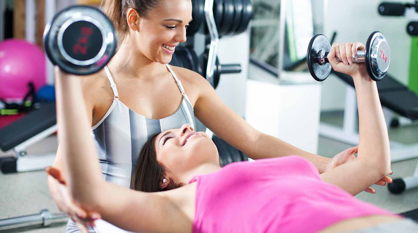 Trainerin unterstützt Frau beim Hanteltraining auf einer Bank im Fitnessstudio.