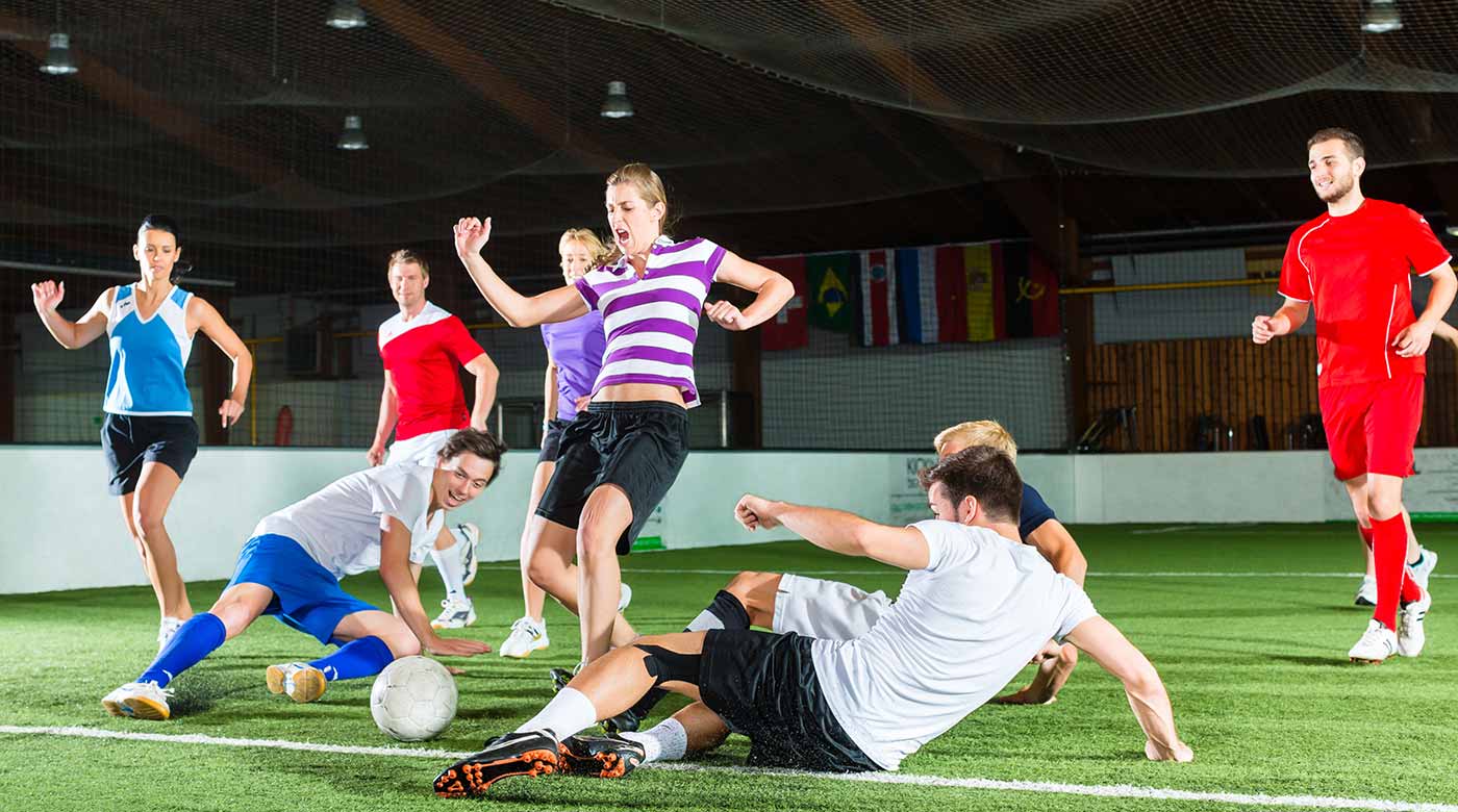 Menschen spielen Fußball in einer Indoor-Halle mit Flaggen im Hintergrund.