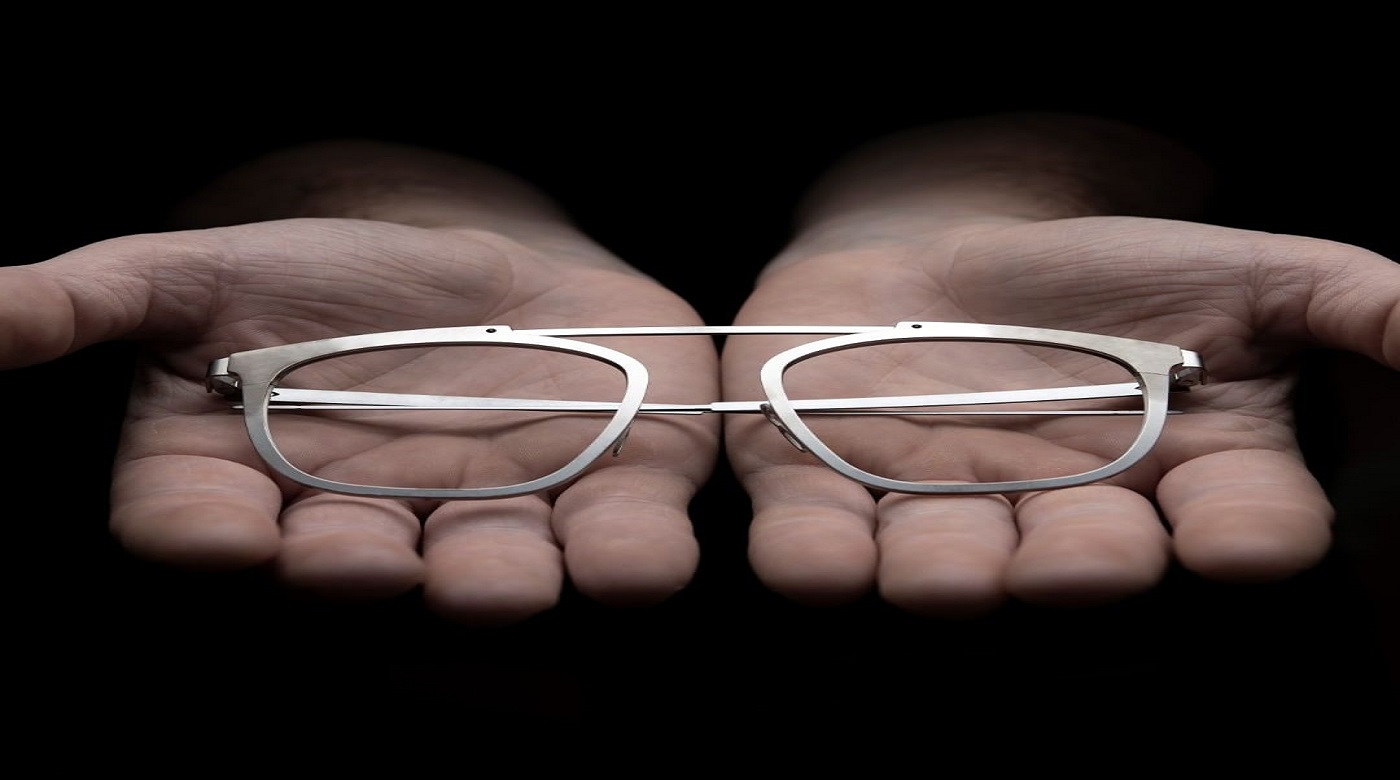 Silberne Brillenfassung wird in zwei offenen Händen vor schwarzem Hintergrund präsentiert.