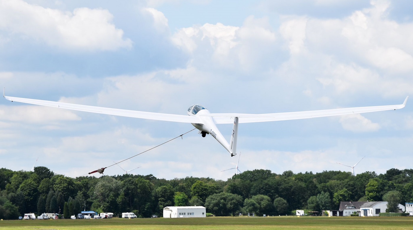 Segelflugzeug im Startvorgang mit Seilwinde, vor blauem Himmel und grüner Landschaft.