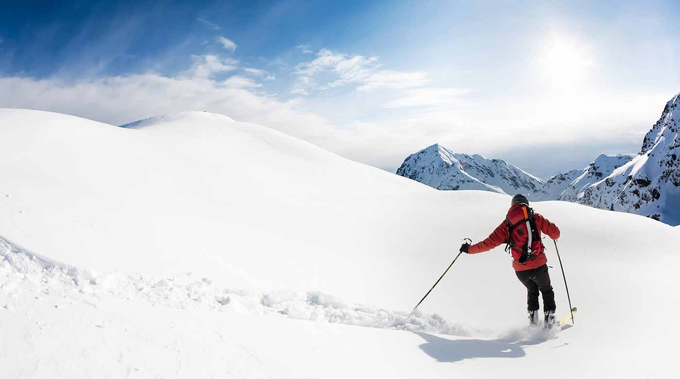 Skifahrer in roter Jacke auf unberührtem Schnee, umgeben von Bergen und blauem Himmel.