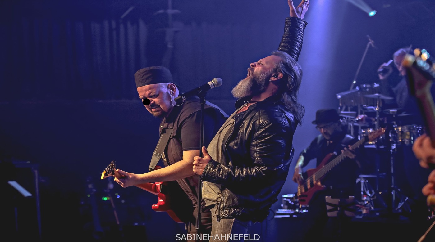 Drei Musiker auf einer Bühne mit Lichteffekten und Instrumenten im Hintergrund.