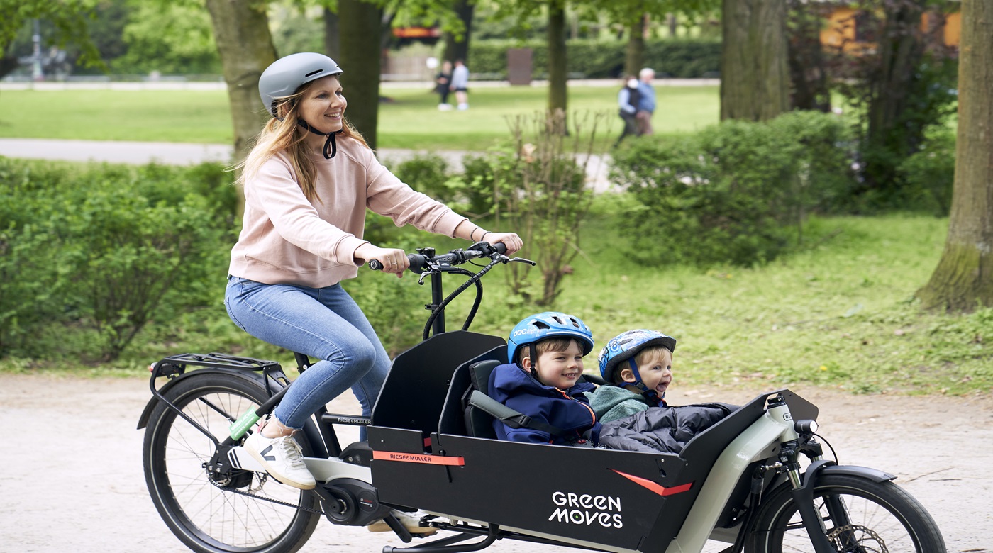 Lächelnde Frau mit Helm mit Lastenrad und 2 Kindern mit Helm