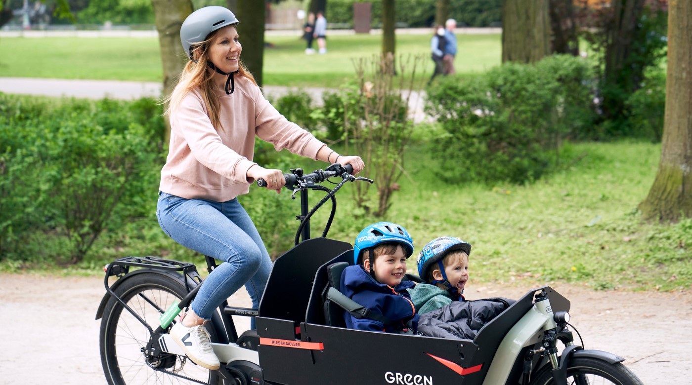Lächelnde Frau mit Helm mit Lastenrad und 2 Kindern mit Helm